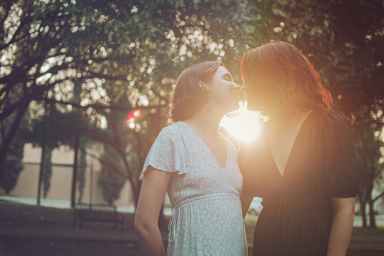 Twee vrouwen op de voorgrond geven elkaar een kus, de zon schijnt achter ze en er zijn bomen te zien op de achtergrond.