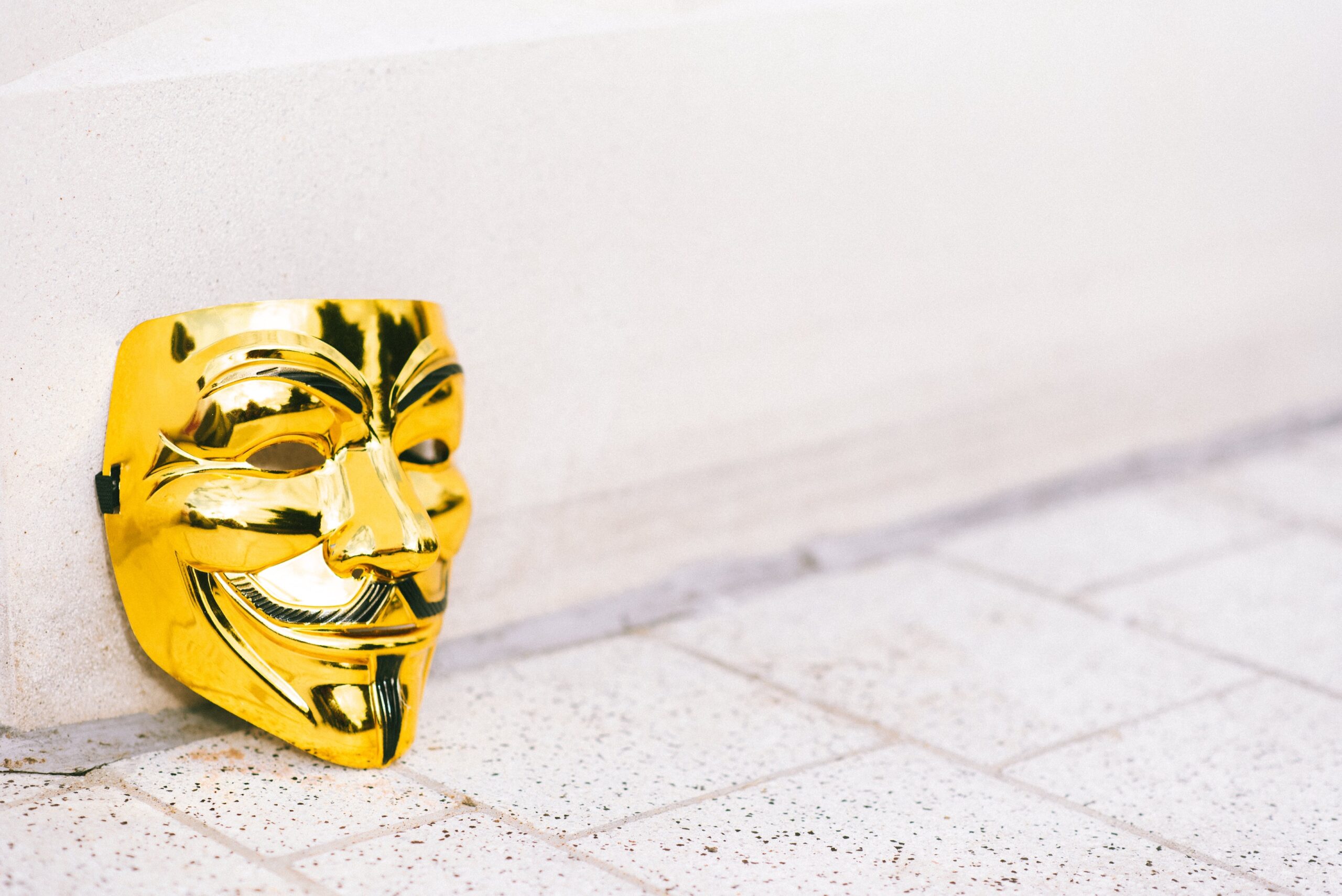 Een gouden masker op een beige vloer rustend tegen een muur van gebroken wit, alsof iemand het masker net heeft afgedaan en op de grond heeft gezet