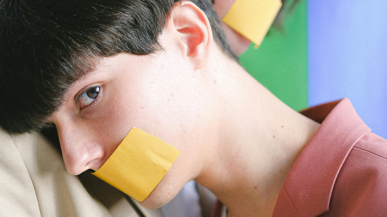 Persoon waarvan mond afgeplakt is met tape, leunt op schouder van een ander persoon met op achtergrond een regenboogvlag